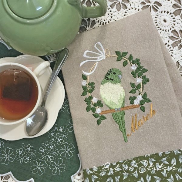 Embroidered tea towel with March Parakeet perched on swing with leafy vine and flowers - machine embroidery design by A Bit of Stitch
